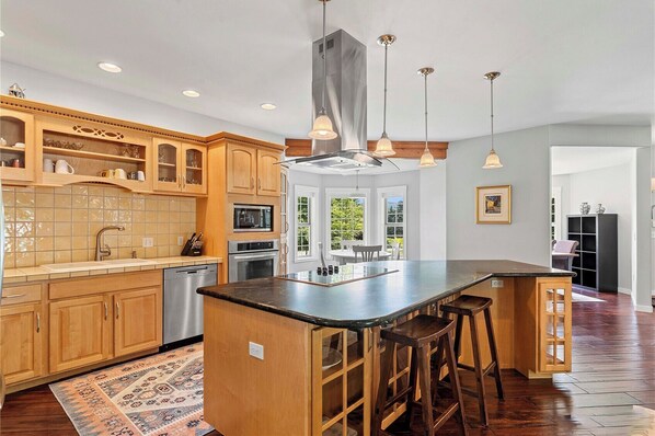 A modern kitchen with wooden cabinets, a central island with stools, a built-in oven, and pendant lighting.