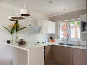 A modern kitchen with white cabinetry, a central island, pendant lights, and a window with a view of palm trees. - Superb upscale villa in Grand Baie in a gated community (Grand Baie)
