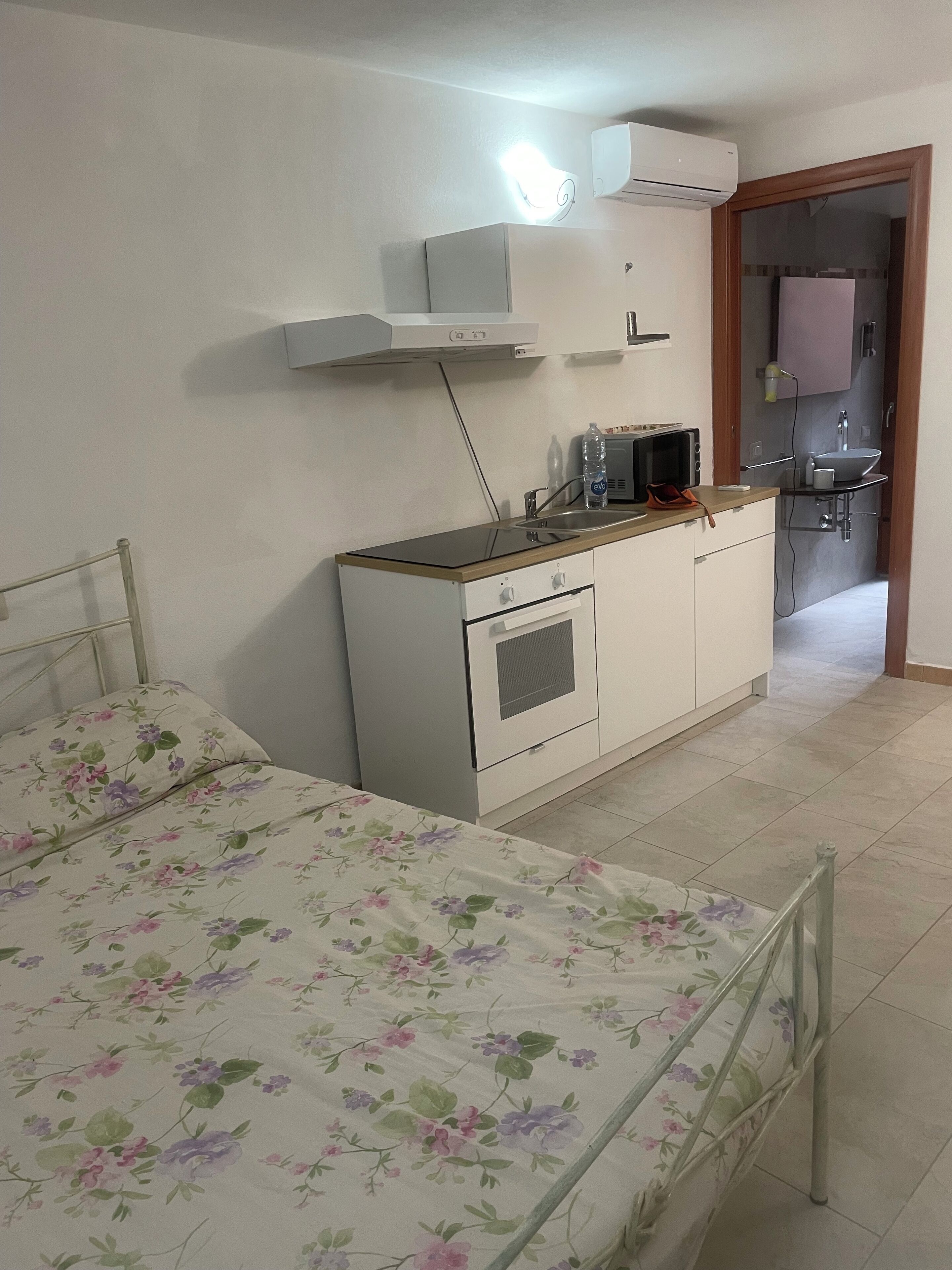 Ein Schlafzimmer mit Bett, einer kleinen Küchenzeile und einem durchgehenden Türspalt sichtbaren Badezimmer.