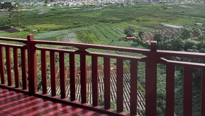Un balcon rouge offrant une vue sur des champs en terrasses et des montagnes lointaines, sous un ciel partiellement nuageux.