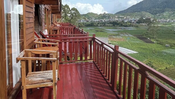 Une terrasse en bois, dotée d’un plancher rouge et d’une balustrade, qui offre une vue sur un paysage verdoyant composé de champs et de collines lointaines.