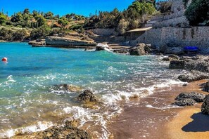 A coastal beach with clear turquoise water, rocky shoreline, and a stone wall. - Villa Artemis in Small Kounoupi Porto Heli (Porto Cheli)