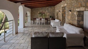 Patio esterno con pareti in pietra, soffitto in legno, un divano bianco, poltrone in vimini e un tavolo con un centrotavola floreale.