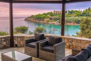 A covered outdoor seating area with wicker furniture and a glass table, overlooking a coastal landscape with a house in the background. - Villa Artemis in Small Kounoupi Porto Heli (Porto Cheli)