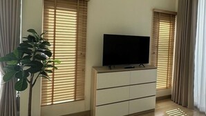 A living room with a flat-screen TV on a white cabinet, large windows with blinds, a potted plant, and a glass coffee table. - HOUSE NO.89 (Chiang Mai)
