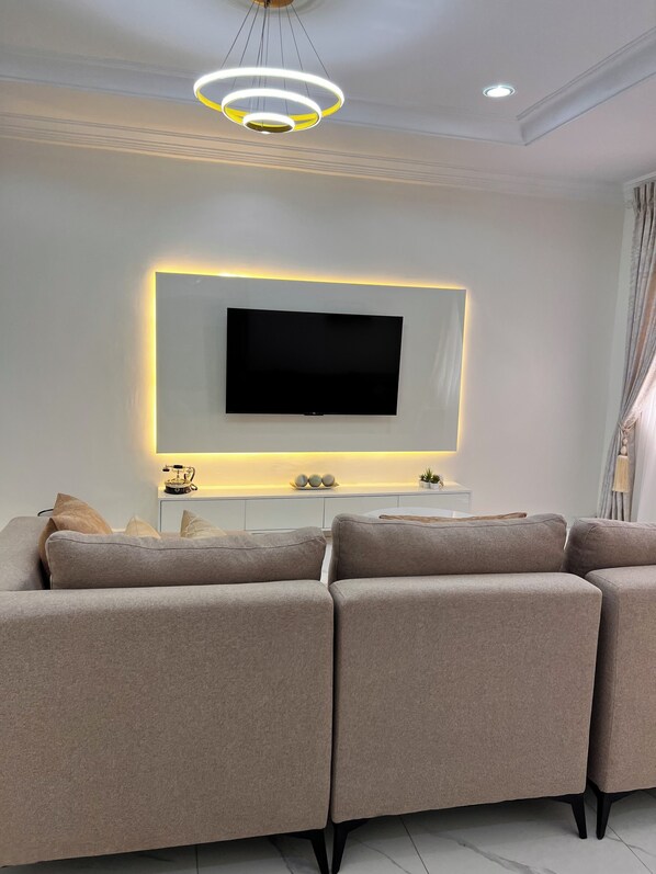 Ett modernt vardagsrum med en stor, platt-TV som är monterad på en vit vägg, en beige hörnsoffa, ett litet vitt konsolbord med dekorativa föremål och en taklampa.