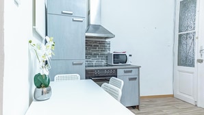 Una cocina compacta con microondas, estufa y un respaldo de ladrillo. Hay una mesa blanca con dos sillas y una planta en maceta.