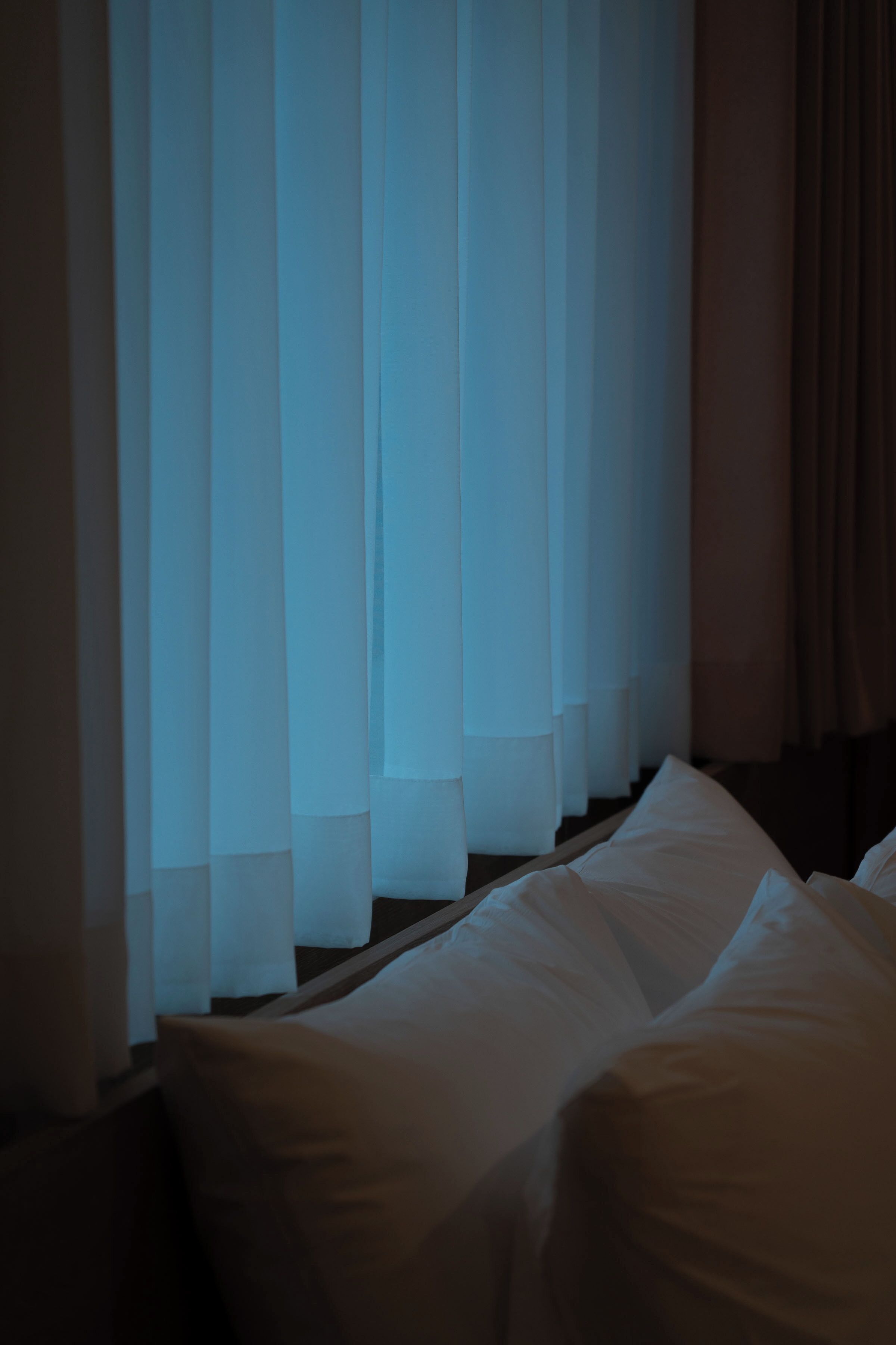 A hotel room with a bed, white curtains, and a window with blinds.