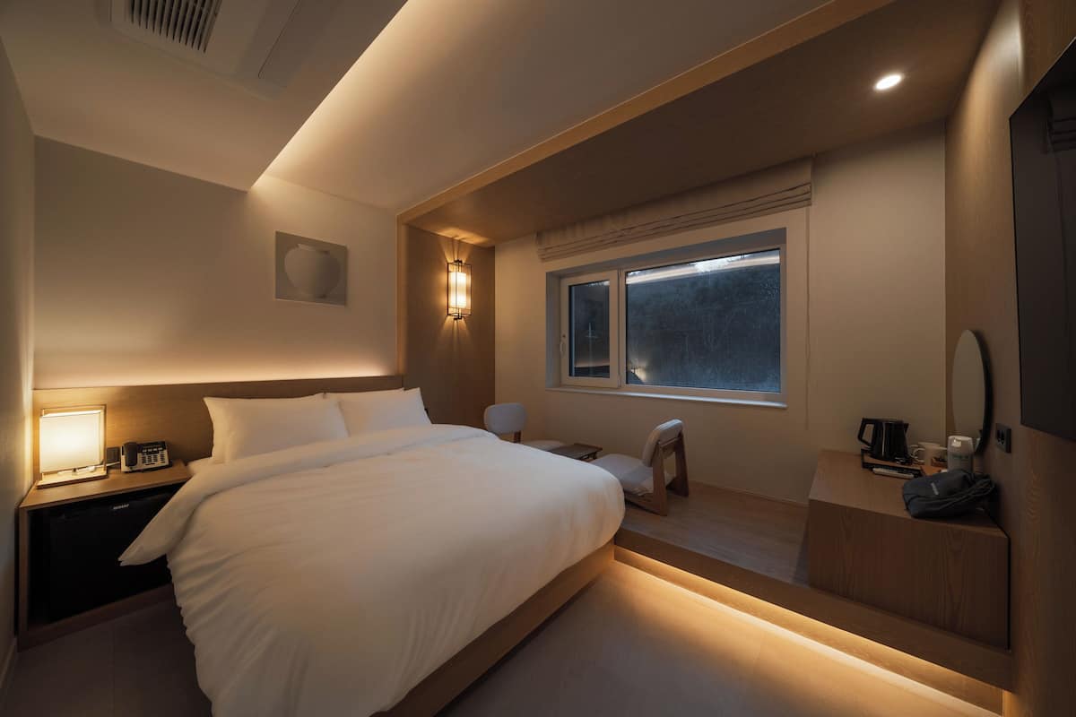 A modern hotel room with a large bed, bedside table with a lamp, a small desk with a chair, a window with a view, and a mirror on the wall.