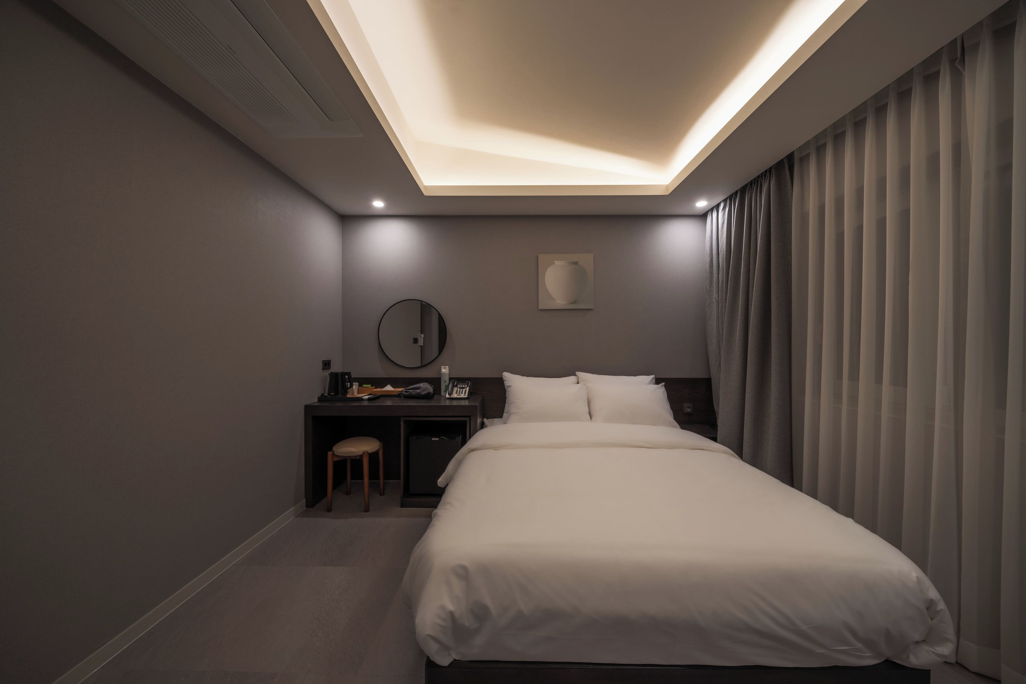 A modern hotel room with a large bed, a desk with a chair, a small table, and a wall-mounted mirror.