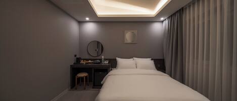 A modern hotel room with a large bed, a desk with a chair, a small table, and a wall-mounted mirror.