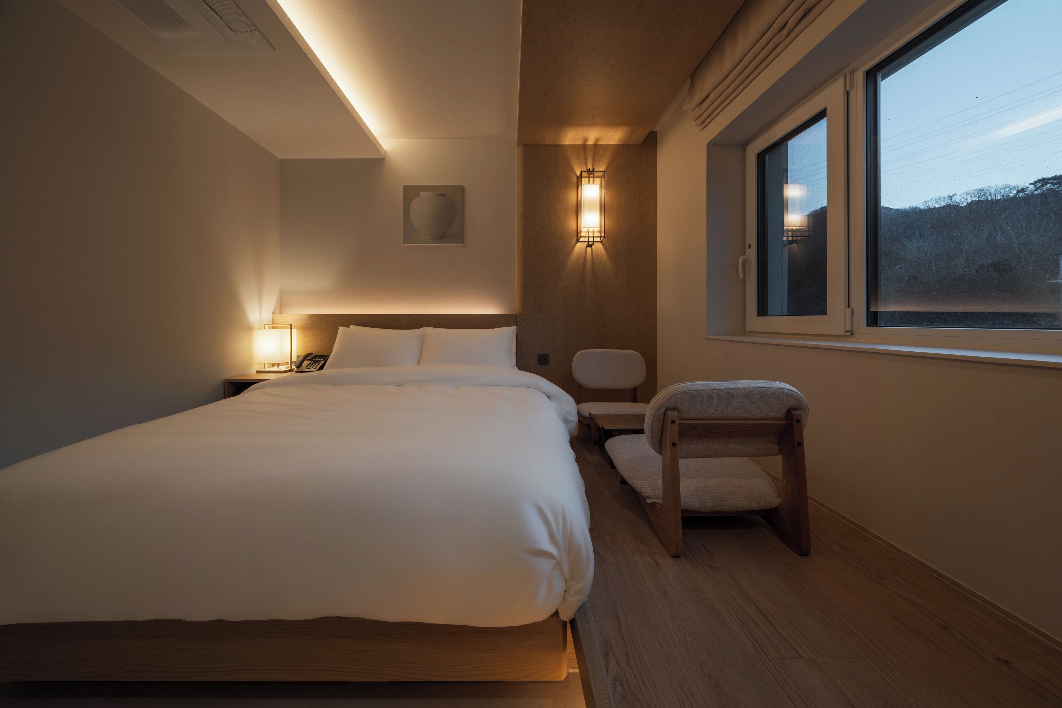 A modern hotel room with a large bed, a nightstand, a wall-mounted lamp, a wooden chair, and a window with a view of the outdoors.