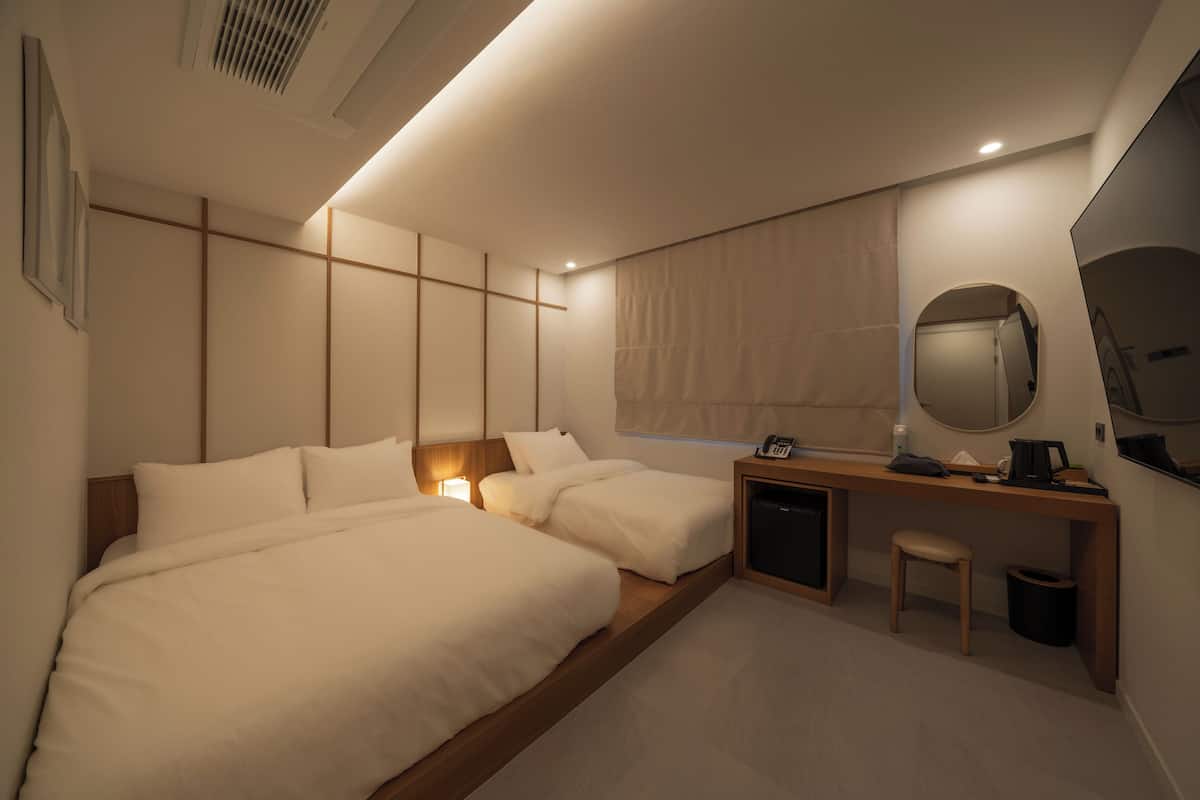 A modern hotel room with two beds, a desk, a chair, a small table, a mirror, and a window with curtains.