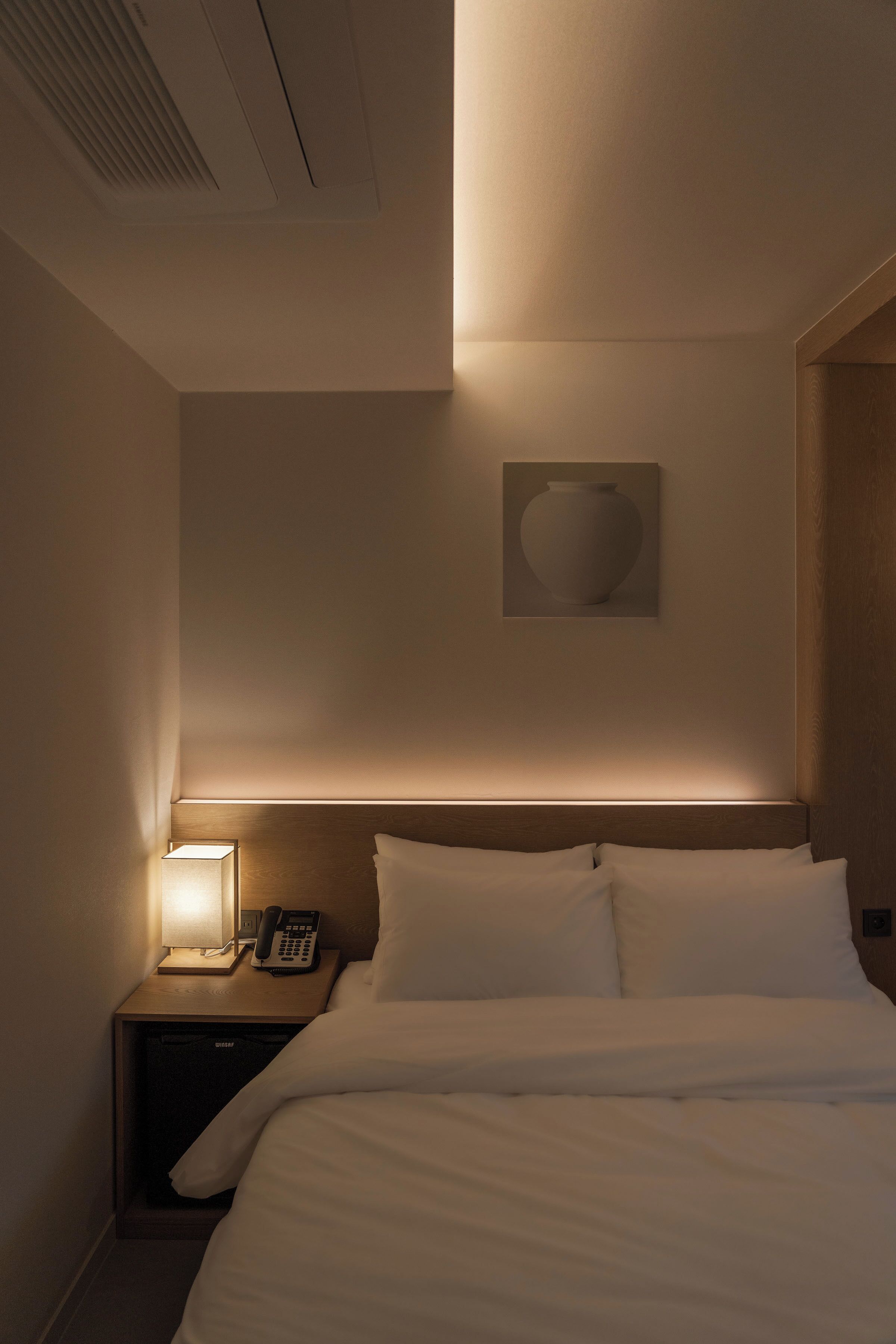 Habitación de hotel con una cama, una mesita de noche con lámpara, un teléfono y una obra de arte colgada en la pared.