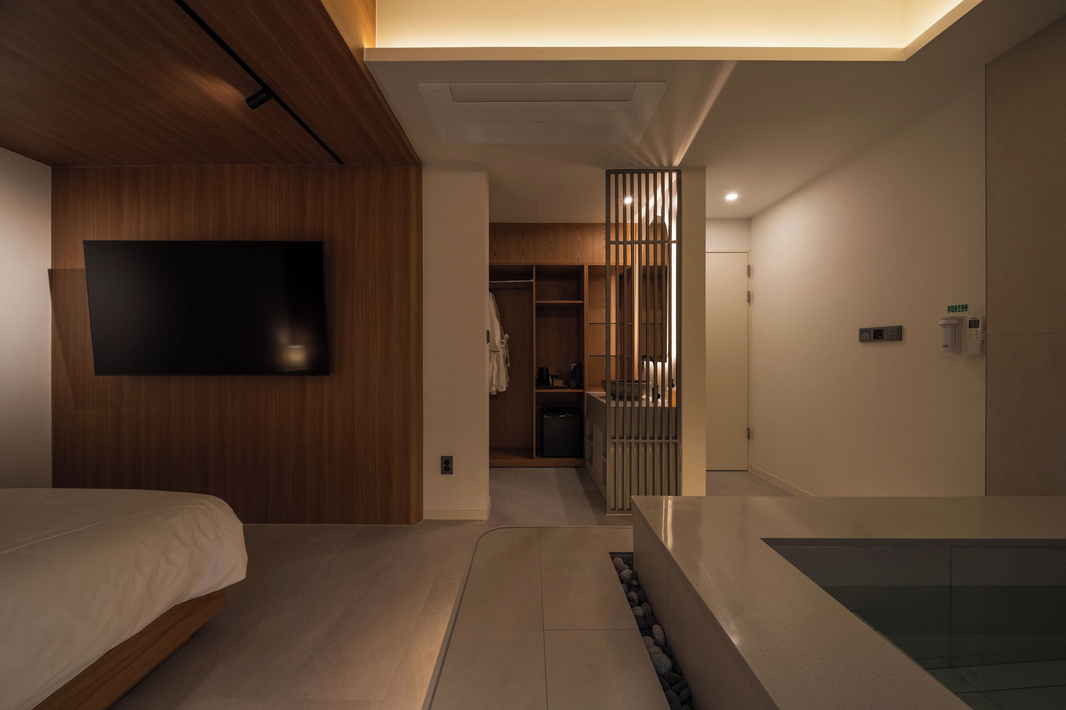 A modern hotel room with a wooden ceiling, a flat-screen TV mounted on the wall, a bed, a small bathroom with a glass shower, and a walk-in closet.