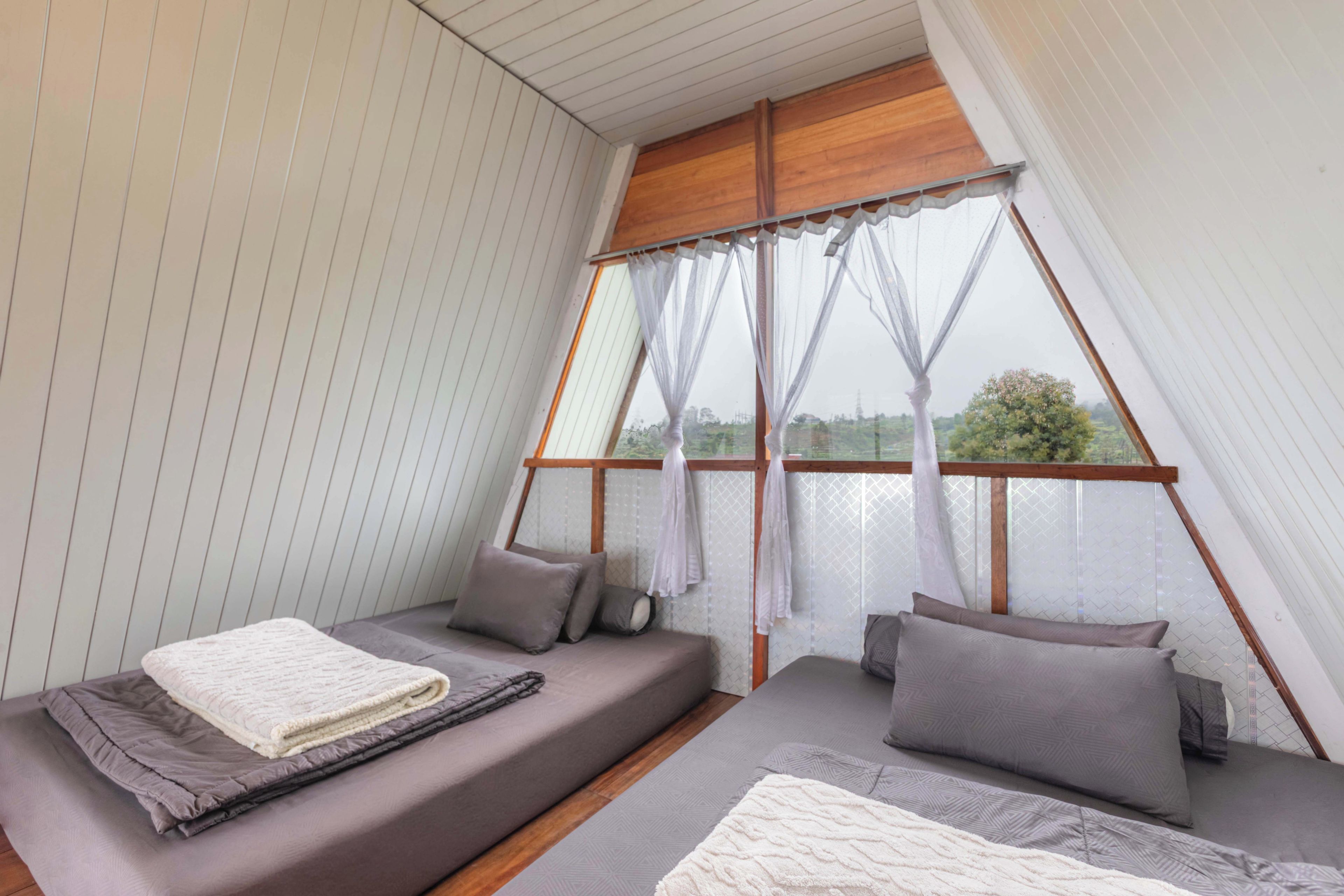 A room with a sloped ceiling, a bed with a white blanket, two pillows, and a view through a window with sheer curtains.