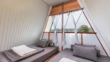 A room with a sloped ceiling, a bed with a white blanket, two pillows, and a view through a window with sheer curtains.