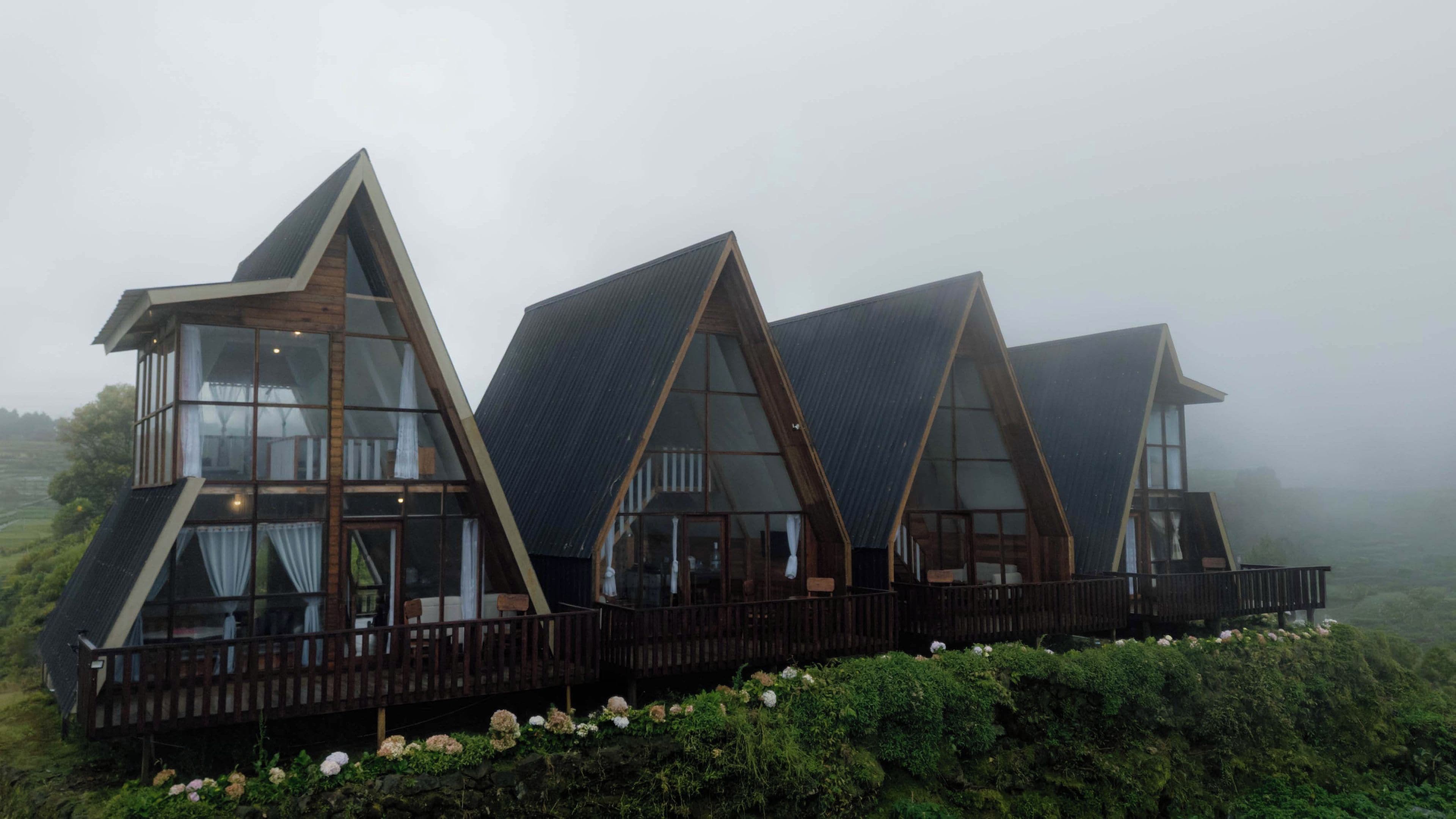 A row of triangular-roofed cabins with large windows and wooden balconies, set in a lush, green landscape.