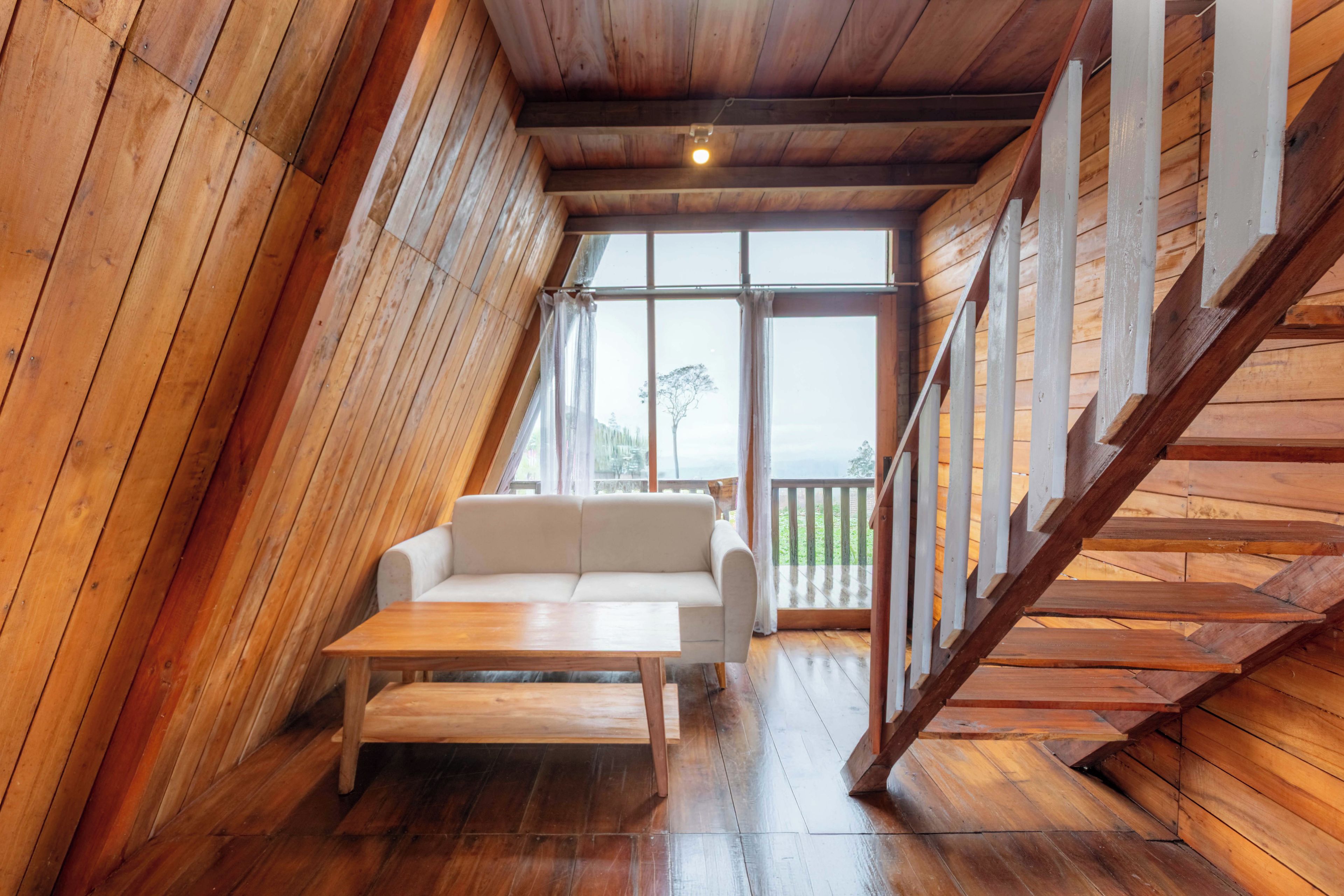 A loft-style room with a wooden sloped ceiling, a white sofa, a wooden table, and a large window offering a view of the outdoors.