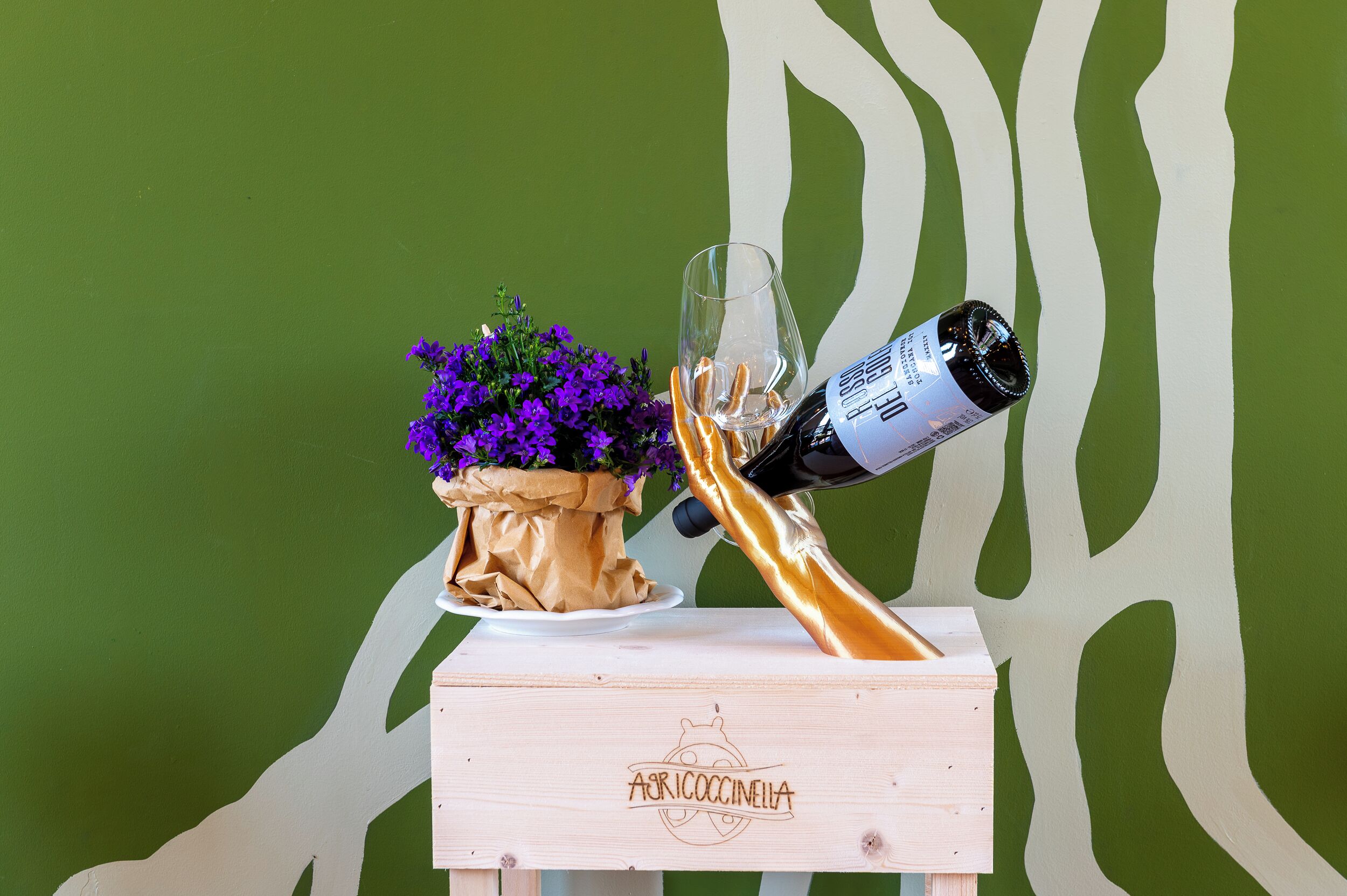 Una caja de madera con la inscripción 'Apericoccinella', una copa de vino blanco y una planta en maceta con flores moradas.