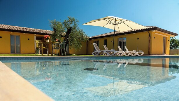 Una piscina cristalina con sillones de descanso y una sombrilla, un edificio amarillo con techo de tejas y un árbol.