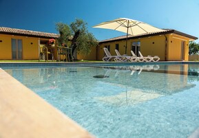 Una piscina cristalina con sillones de descanso y una sombrilla, un edificio amarillo con techo de tejas y un árbol.