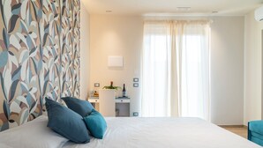 Una cama bien hecha con almohadas azules, un escritorio con una planta y una ventana con cortinas translúcidas.