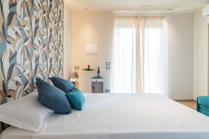 Una cama bien hecha con almohadas azules, un escritorio con una planta y una ventana con cortinas translúcidas.