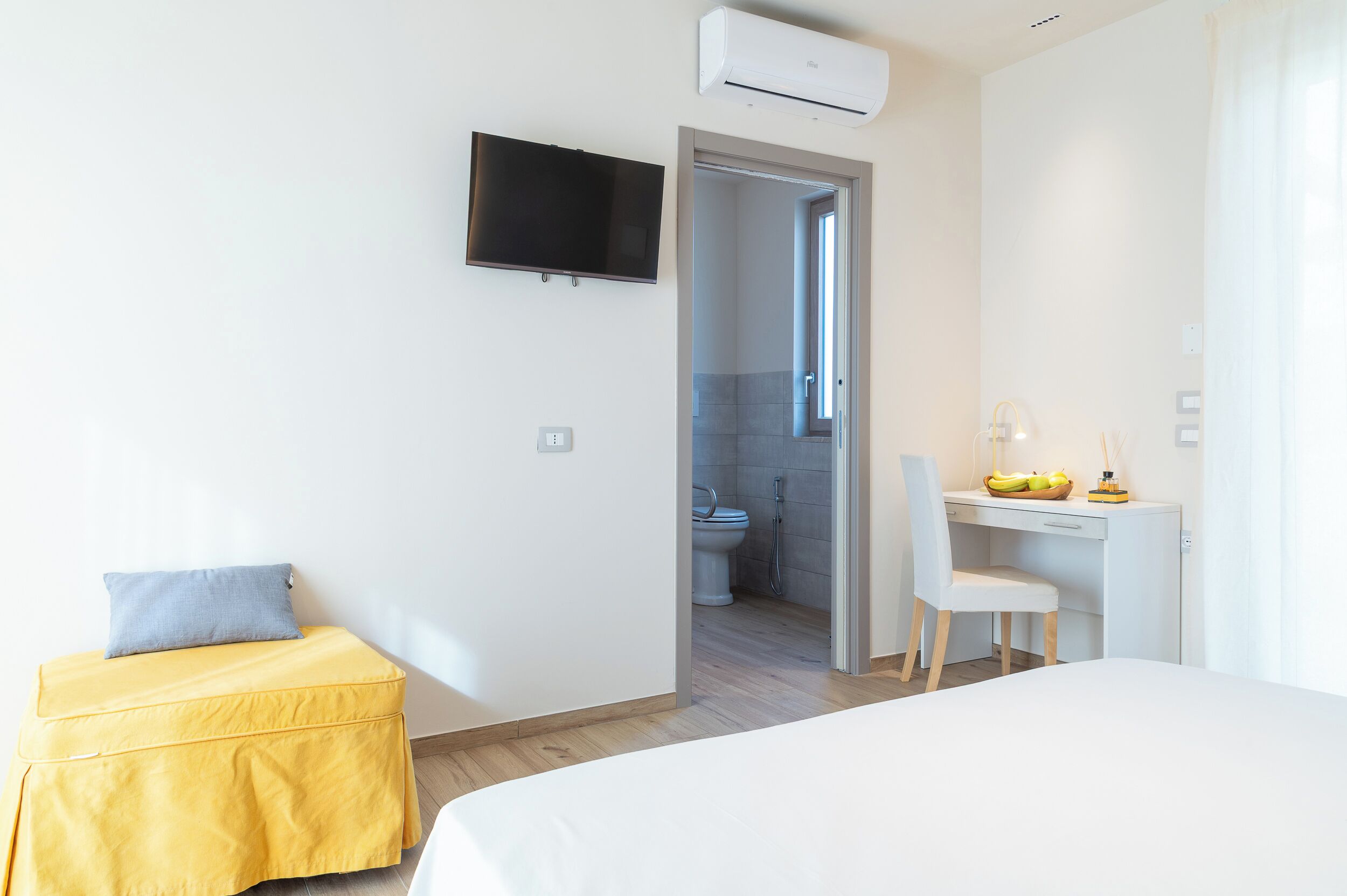 A modern hotel room with a bed, a desk with a chair, a television, and an air conditioning unit.