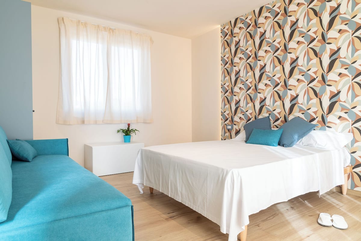 Un dormitorio con cama, sofá, mesita de noche, planta en maceta y pared con estampado.