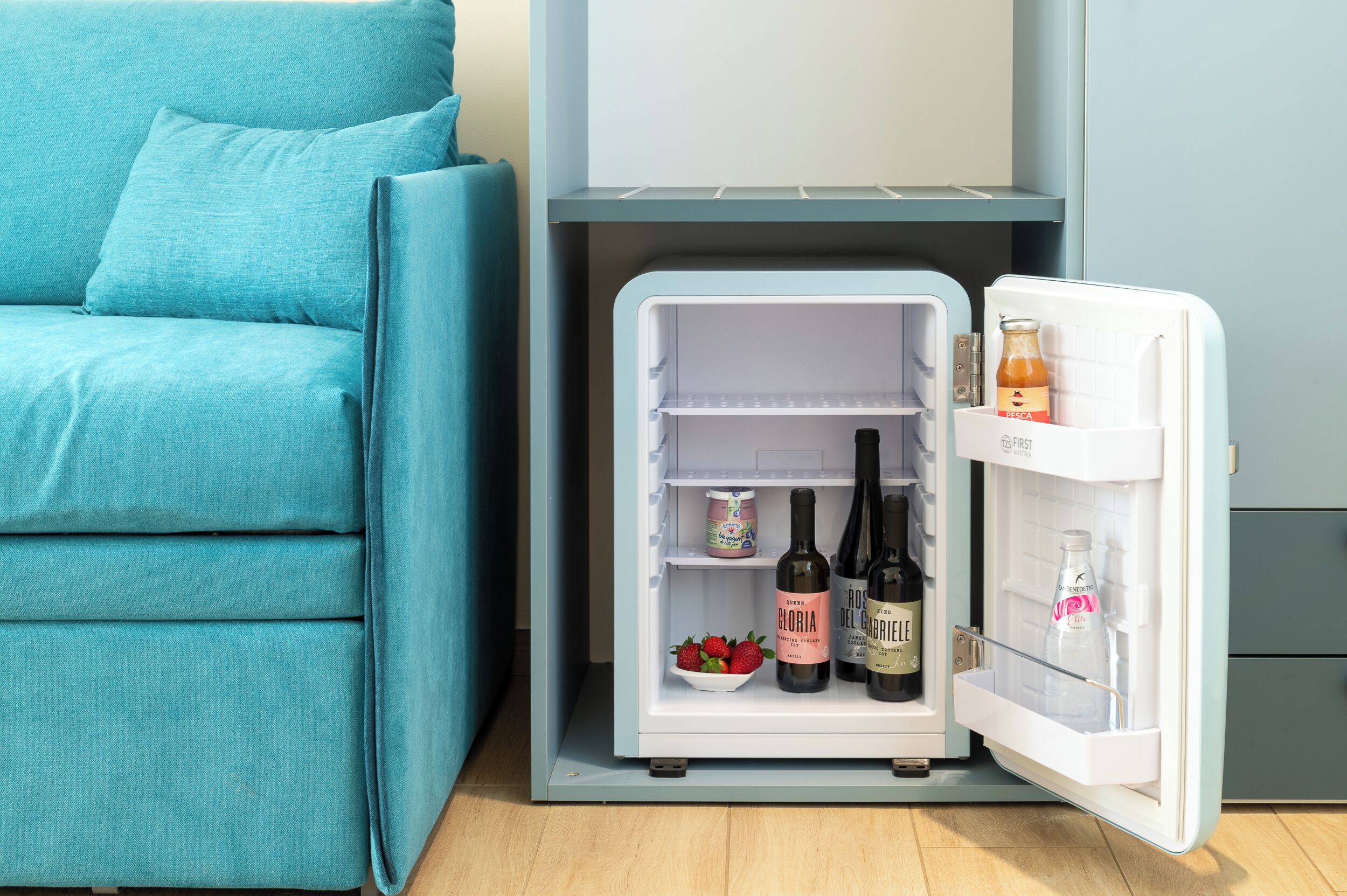 Un refrigerador compacto con una botella de vino rosado, dos botellas de vino tinto, una botella de agua y un tazón de fresas.