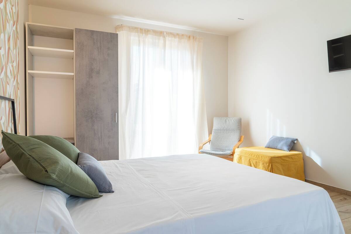 Un dormitorio con cama, una silla, una mesita, un armario y un televisor en la pared.