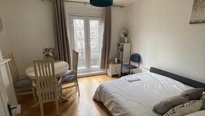 Ein Schlafzimmer mit einem Bett, einem Schreibtisch und einem Stuhl. Es gibt ein Fenster mit Vorhängen, eine Tür und eine Deckenleuchte.