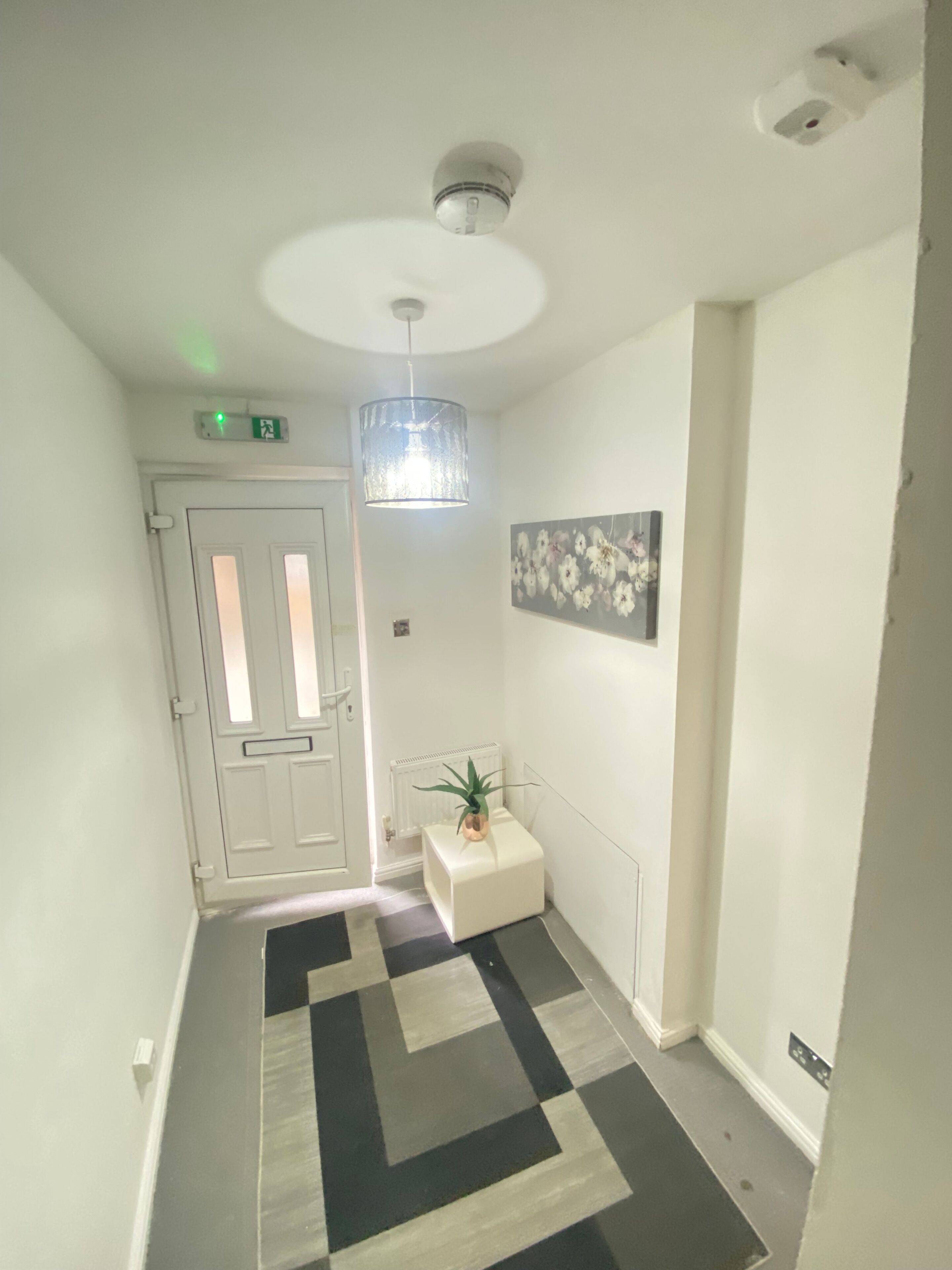 A hallway with a patterned floor, a white door, a radiator, a small plant, a wall-mounted light fixture, and a ceiling-mounted smoke detector.