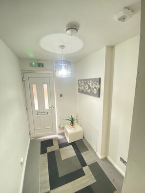 Un corridoio con pavimento decorato, una porta bianca, un termosifone, una piccola pianta, un lampadario a parete e un rilevatore di fumo a soffitto.