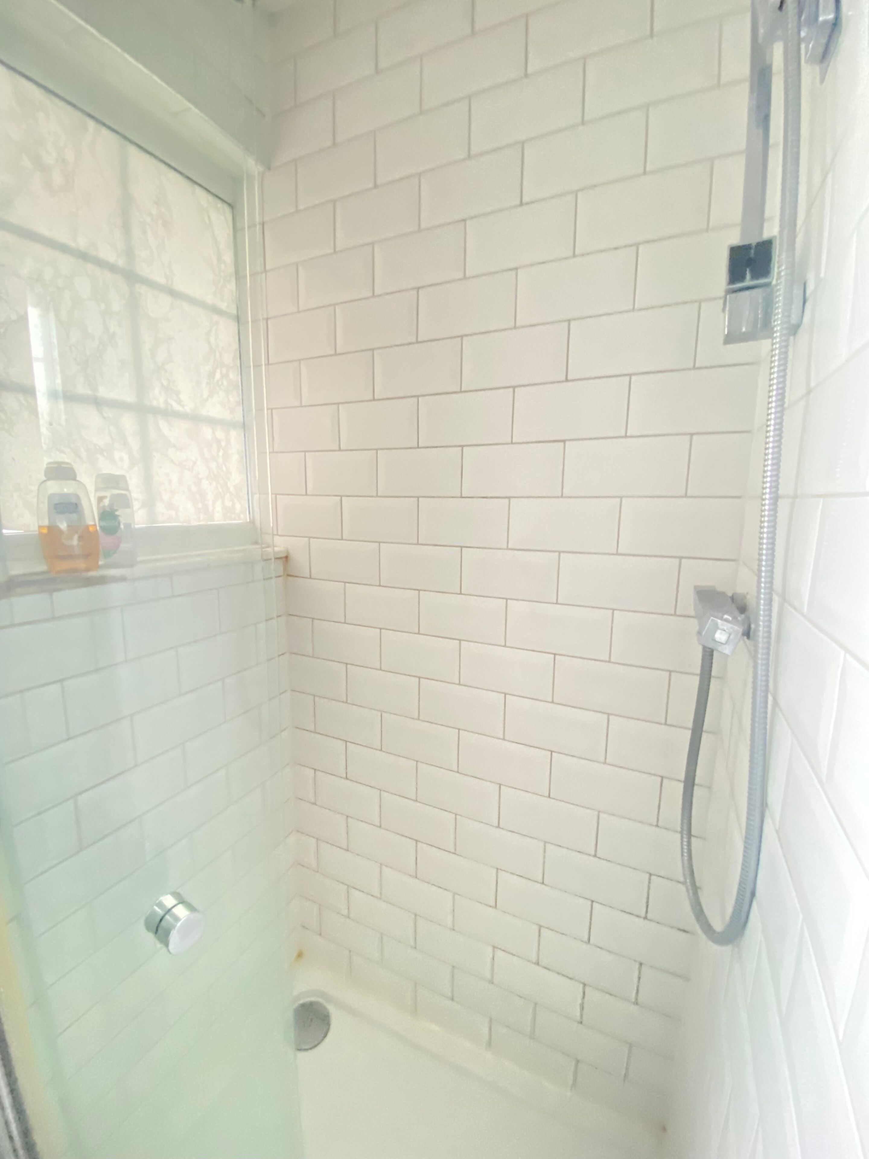 Un espace douche avec des murs recouverts de carreaux blancs et une fenêtre.