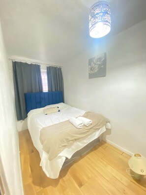 A bedroom with a bed, curtains, a wall-mounted light fixture, and a small table with a lamp. - Luxurious 5 Bedroom Home in Prime London Location (London)