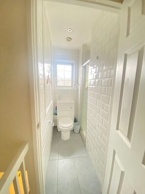 A narrow bathroom with white tiled walls, a toilet, and a window. - Luxurious 5 Bedroom Home in Prime London Location (London)