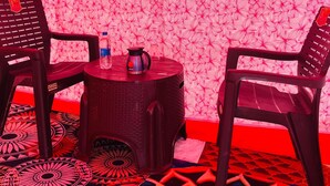 A room with a patterned wallpaper, two black plastic chairs, a small round table with a kettle and a bottle, and a framed picture on the wall. - Prabhu Premi Sevashray (Karchana)