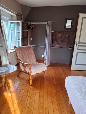 A room with a wooden floor, a wicker chair, a bed, a small table with a doily, a window with curtains, a shower area, and a mirror on the wall. - Stone house 170m³ Pleasant. 2 to 10 people .2 days to 1 or 2 weeks (Tocqueville)