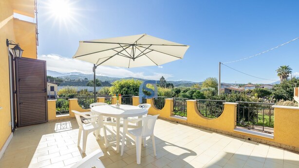 Uma varanda com mesa e cadeiras, um grande guarda-sol e vista para a paisagem.