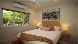 Une chambre à coucher avec un lit, une fenêtre avec des stores, un climatiseur, un ventilateur de plafond, un tableau accroché au mur, des lampes de chevet et un panier en osier posé sur une table de chevet.