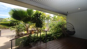 Une terrasse en bois avec une rambarde en métal, entourée d’une végétation tropicale luxuriante et agrémentée d’un fauteuil suspendu.