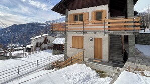 Een chalet met twee verdiepingen, een houten balkon, stenen muren en uitzicht op besneeuwde bergen.