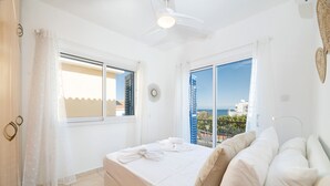 Un dormitorio con cama, cortinas blancas, ventilador de techo y vista al mar.