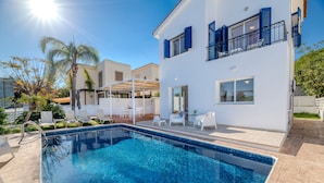 Un área de piscina moderna con una piscina de color azul cristalino, sillas de descanso blancas y un edificio de dos pisos con contraventanas azules.
