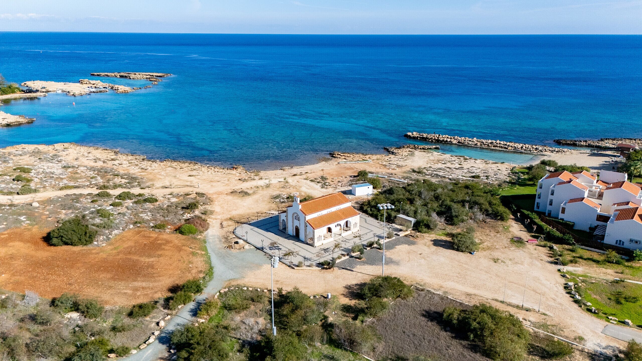 An aerial view of a coastal area with a white building with a red roof, a rocky shoreline, and a clear blue sea.
