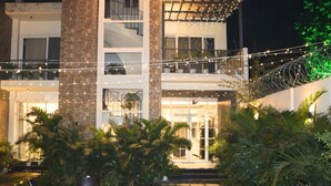 Un edificio de dos pisos con un balcón, luces colgantes y un jardín con sillas y palmeras.