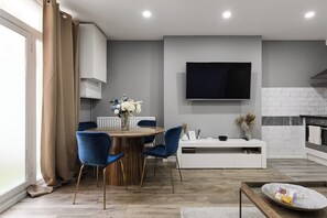Une cuisine moderne équipée d’un téléviseur, de meubles blancs et d’un coin repas avec des chaises bleues et une table en bois.