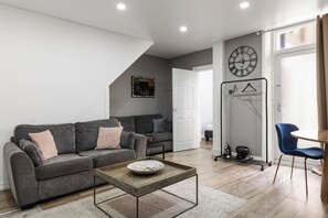 A modern living room with a grey sofa, a coffee table, and a dining area with a blue chair. - 2BR in Kensington, 2 min Earls Court tube (London)